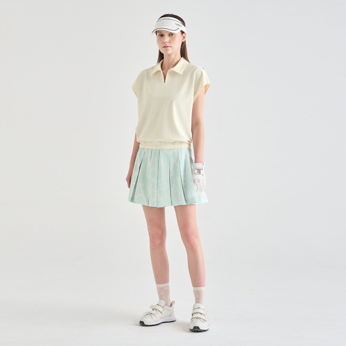 A woman modeling a light yellow sleeveless polo shirt and a light green pleated tennis skirt, paired with a white visor, white glove, white socks, and white sneakers, against a plain light gray studio background.