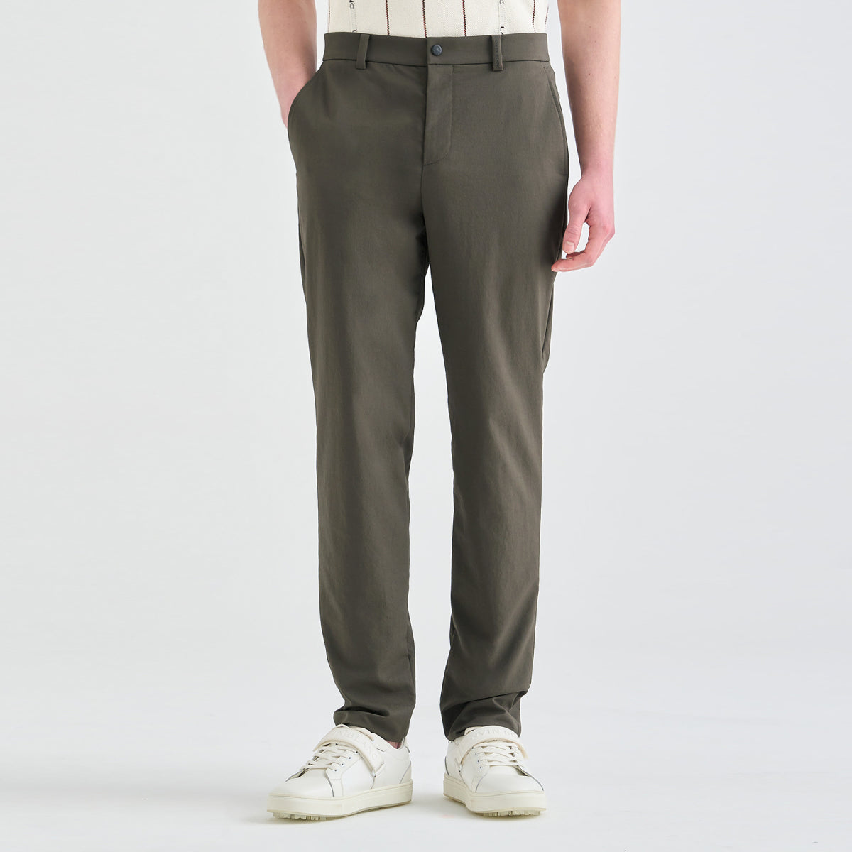 A person wearing slim-fit olive green trousers, styled with a cream and brown striped shirt and white sneakers, shown from the waist down on a light gray studio background.
