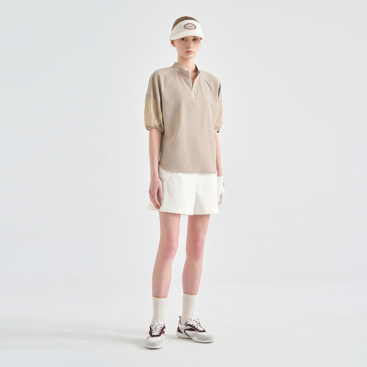 A model stands against a light gray studio background wearing a beige short-sleeve polo-style top, white shorts, white socks, and white sneakers with dark accents. She also wears a white visor.