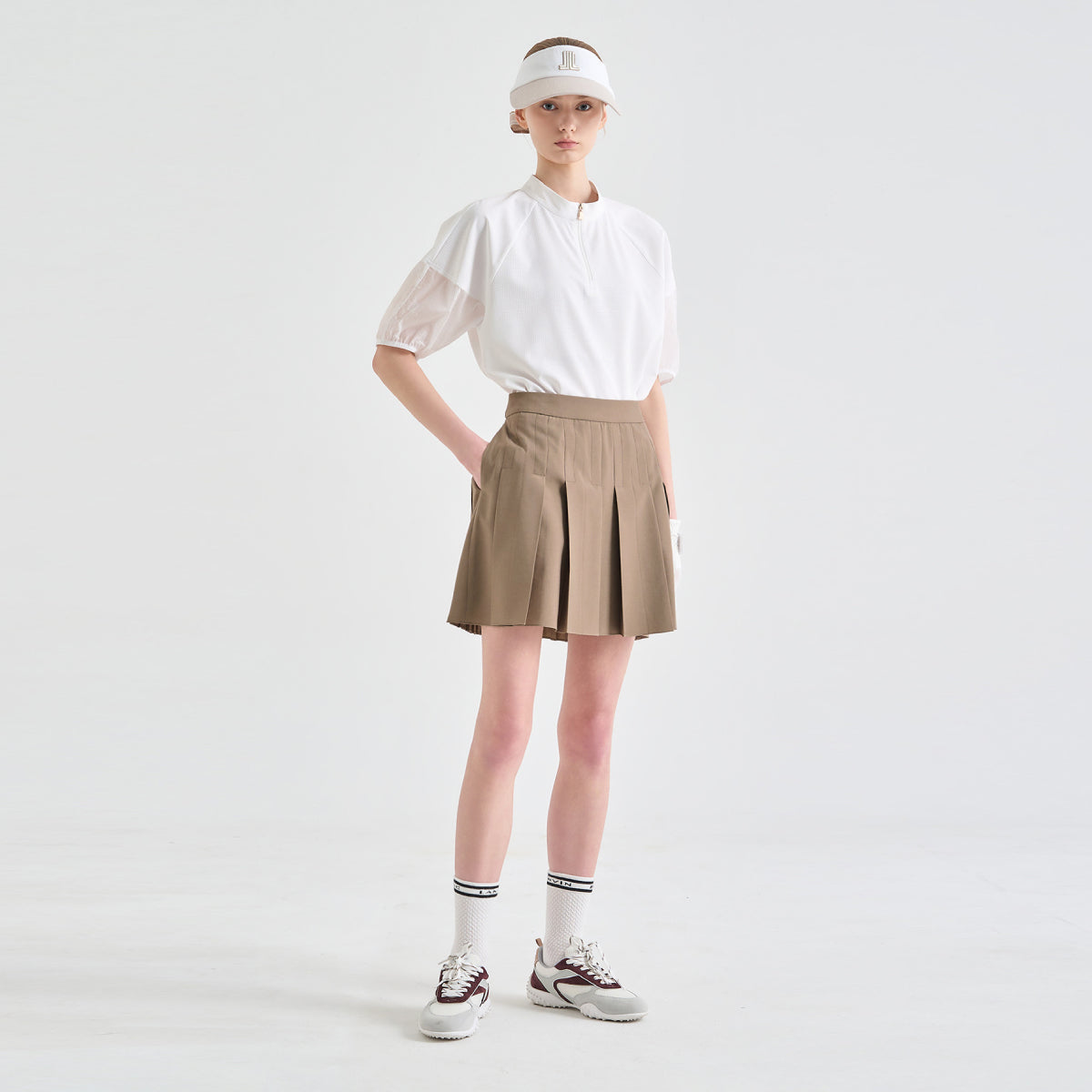 A model stands against a light gray studio background wearing a white short-sleeve blouse with sheer puff sleeves, a beige pleated tennis skirt, a white visor, white socks, and white sneakers.
