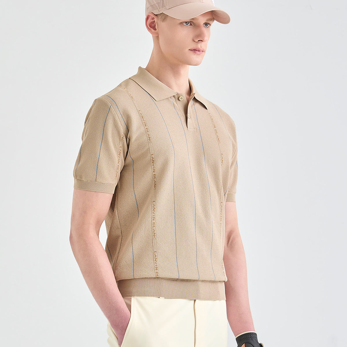A man wearing a beige short-sleeve knit polo shirt with vertical embroidered stripes, paired with cream trousers and a beige cap, photographed against a light gray studio background.