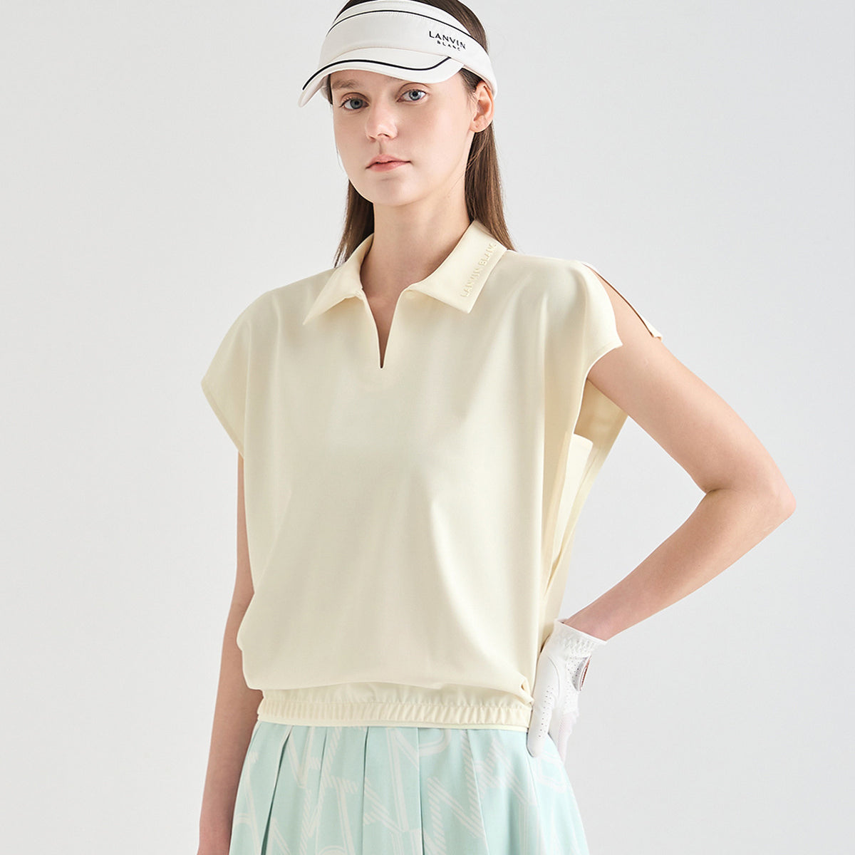 A woman modeling a pale cream short-sleeve polo shirt with a relaxed fit, paired with a white visor and a light mint green patterned skirt, against a solid light gray background.