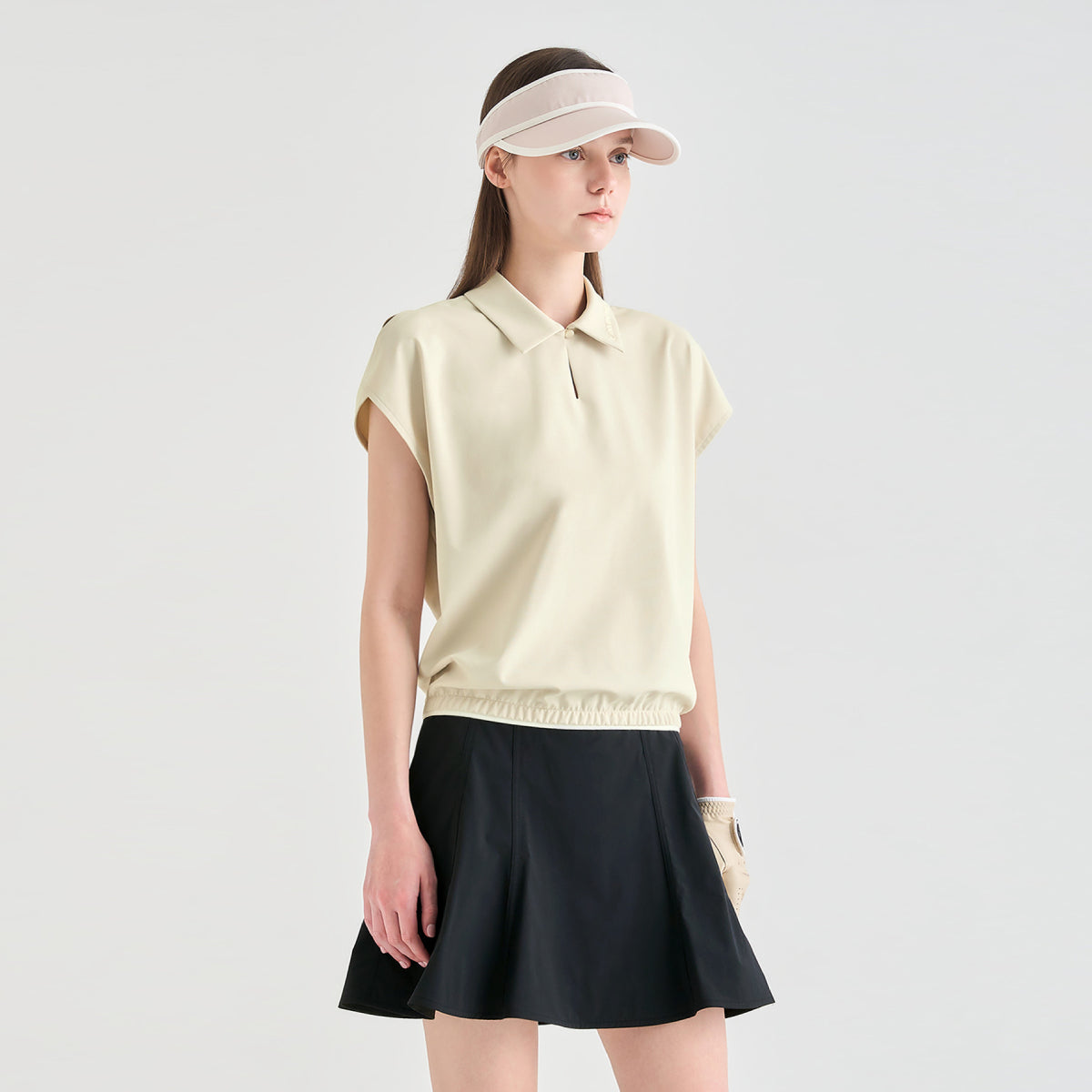 A woman modeling a light cream short-sleeve polo shirt with a black pleated tennis skirt and a beige visor, standing against a solid light gray background.