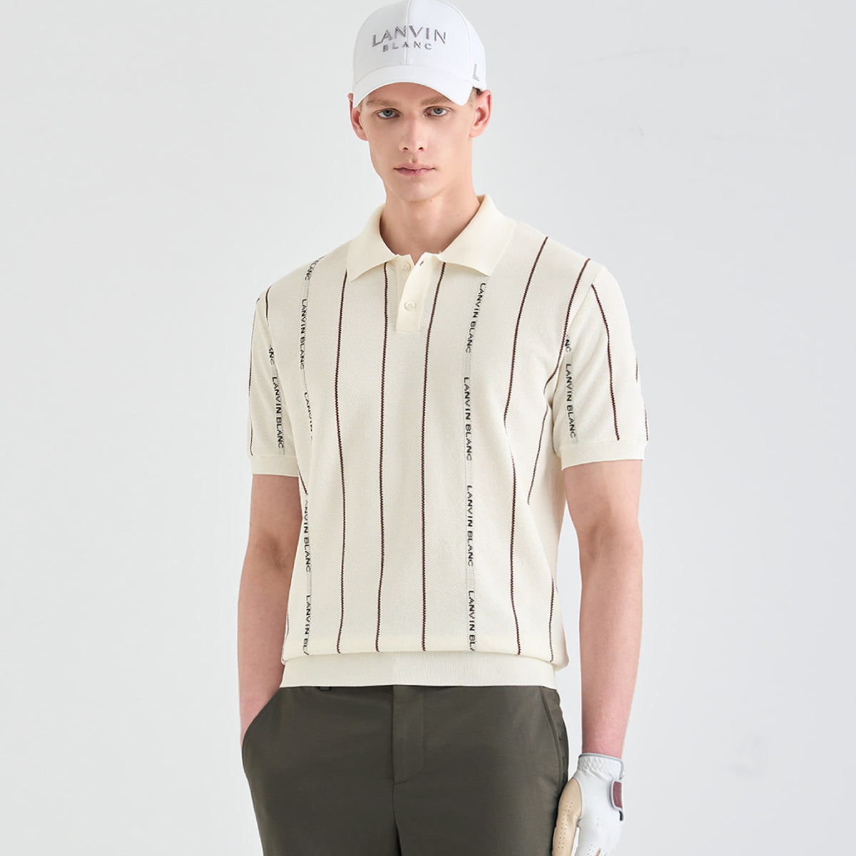 A male model wearing a cream-colored short-sleeve polo shirt with vertical brown stripes and 'LANVIN BLANC' text, paired with dark olive trousers and a white cap.