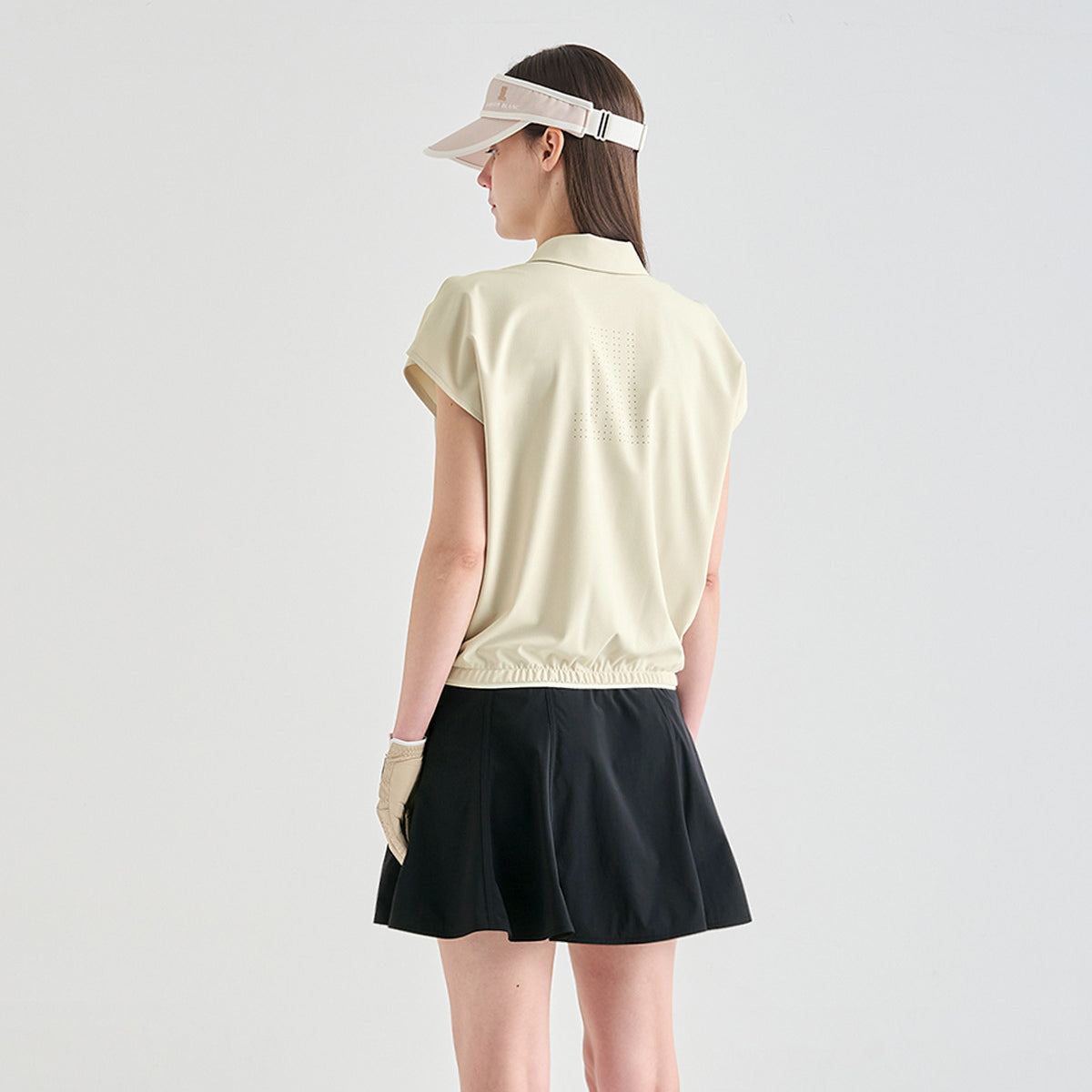 A woman shown from the back wearing a light beige short-sleeve collared athletic top, a black skirt, a beige visor, and beige gloves, against a plain light gray studio background.