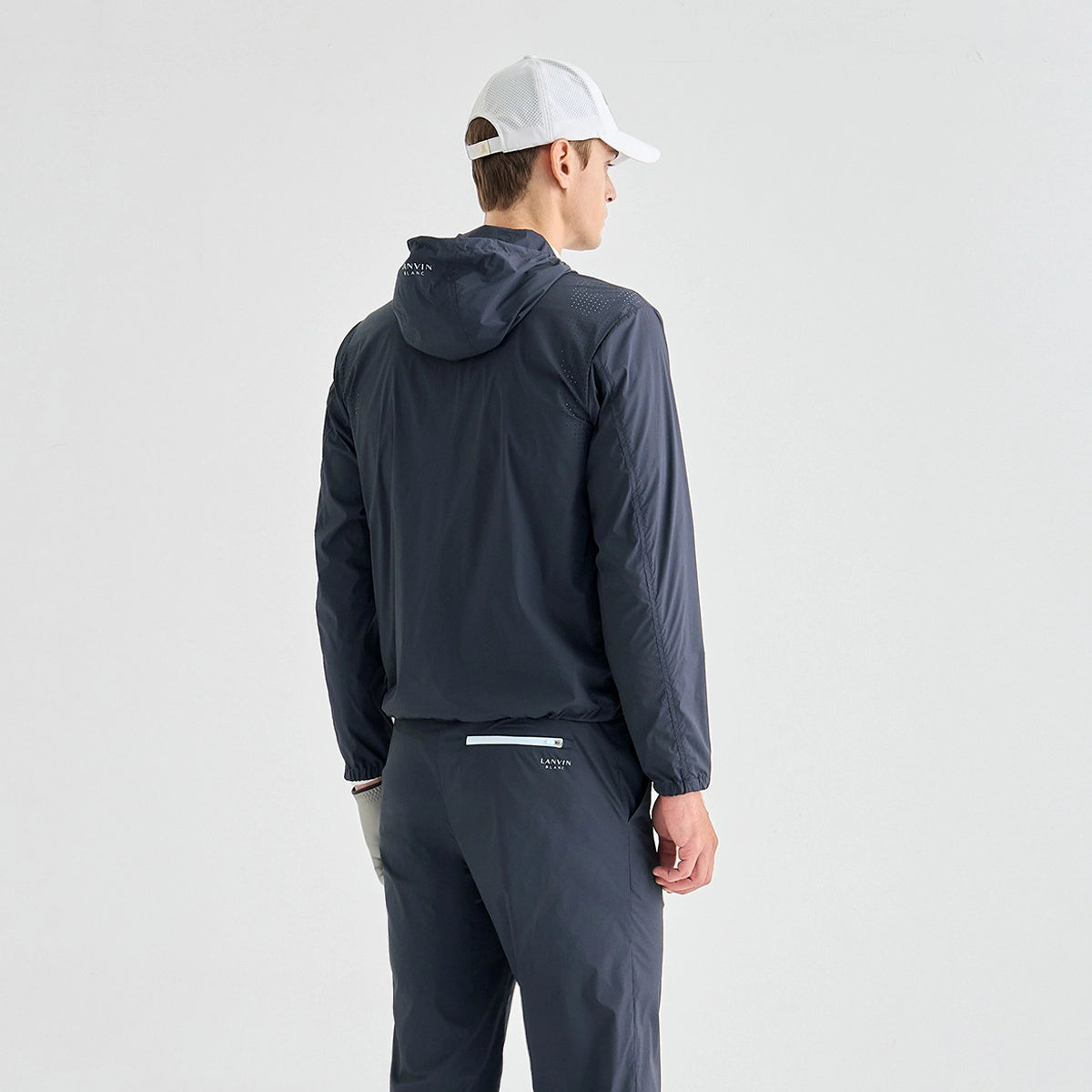 A man wearing a dark navy hooded jacket and matching pants, viewed from the back, wearing a white cap against a light gray studio background.