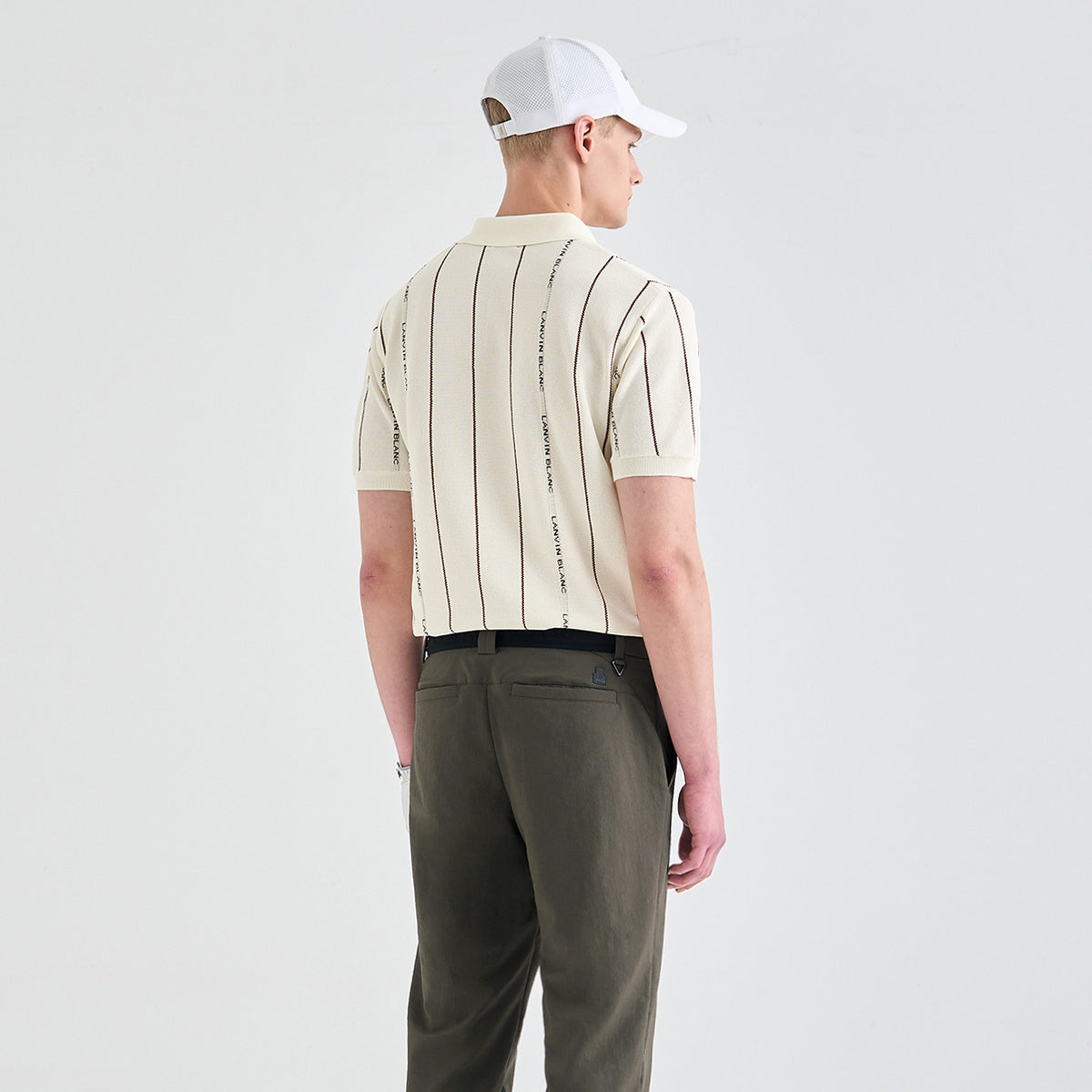 A man is shown from behind wearing a cream short-sleeve polo shirt with thin vertical black stripes and black text running down the center, paired with olive green trousers and a white baseball cap, against a light gray studio background.
