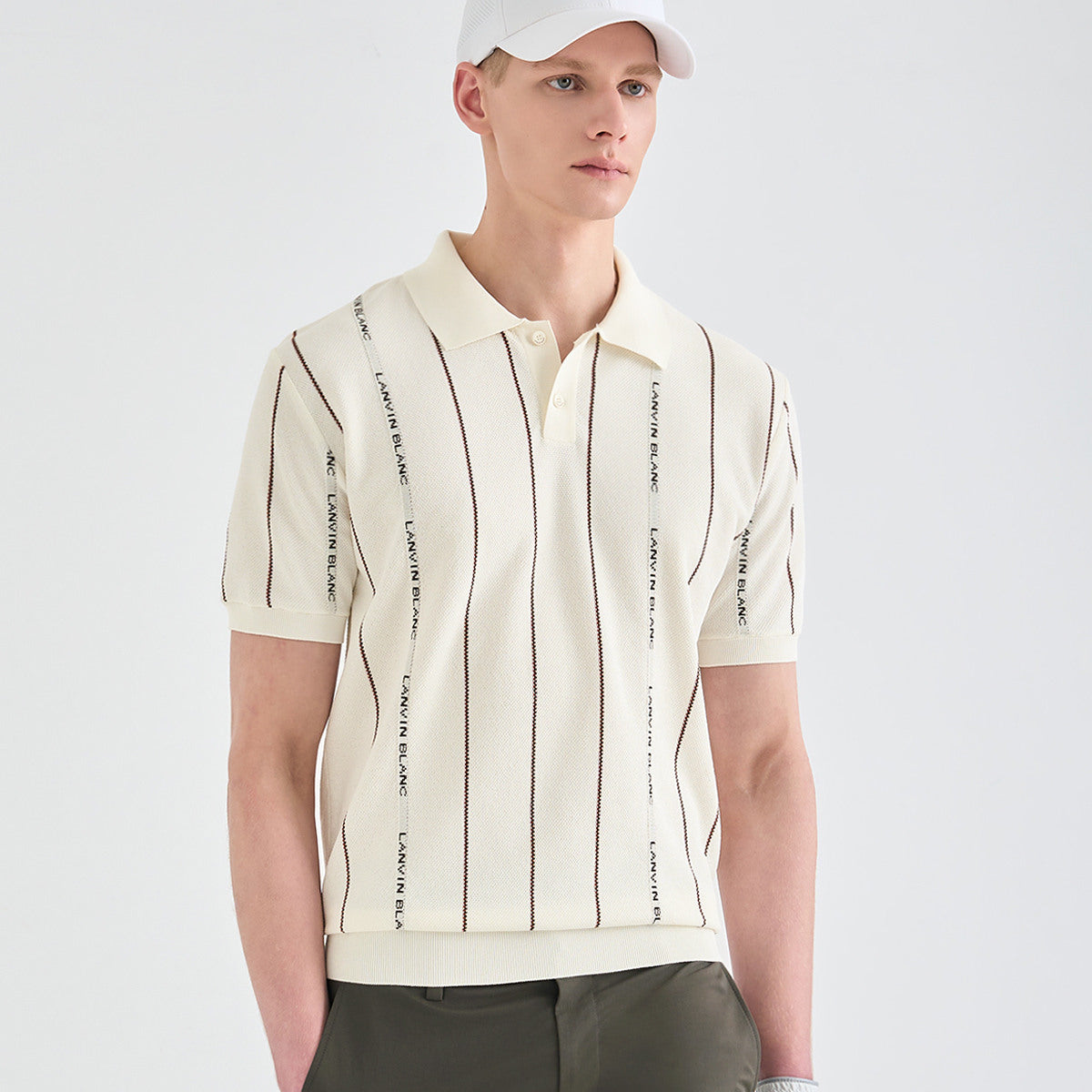A man wearing a cream-colored short-sleeve polo shirt with thin vertical dark stripes and black text running down the sides, paired with dark trousers and a white cap, against a light gray studio background.