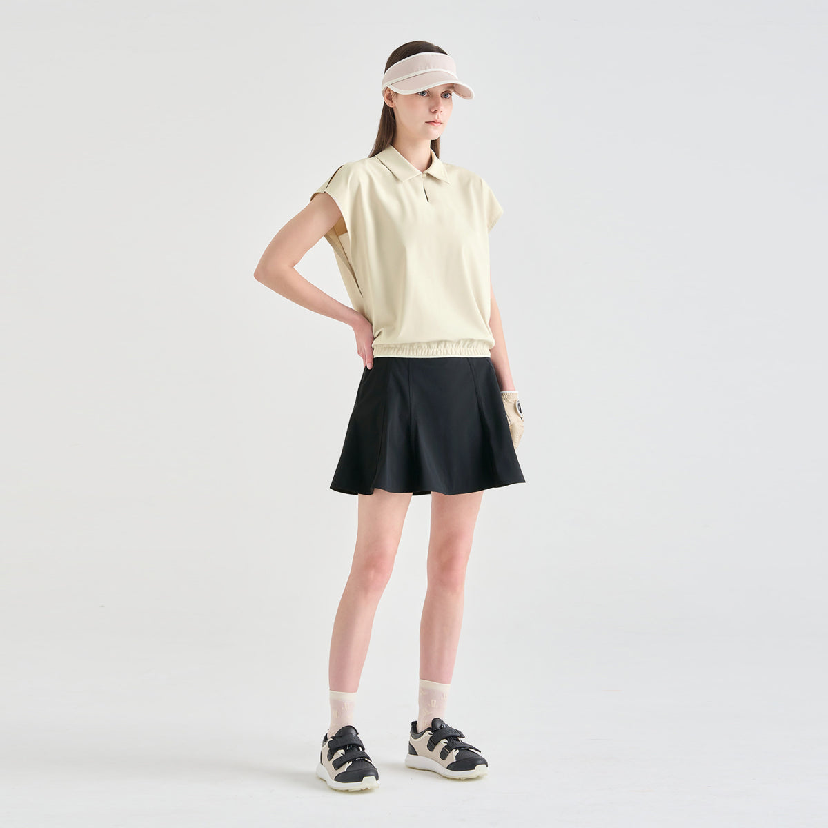 A model stands in a studio wearing a light cream sleeveless polo shirt, a black athletic skirt, a matching visor, white socks, and black-and-white sneakers against a light gray background.
