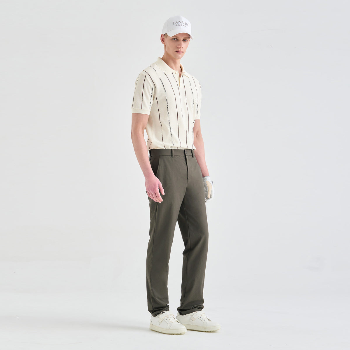 A male model stands in a studio wearing a cream short-sleeve collared shirt with vertical black text, olive green trousers, a white cap, a white golf glove, and white sneakers.