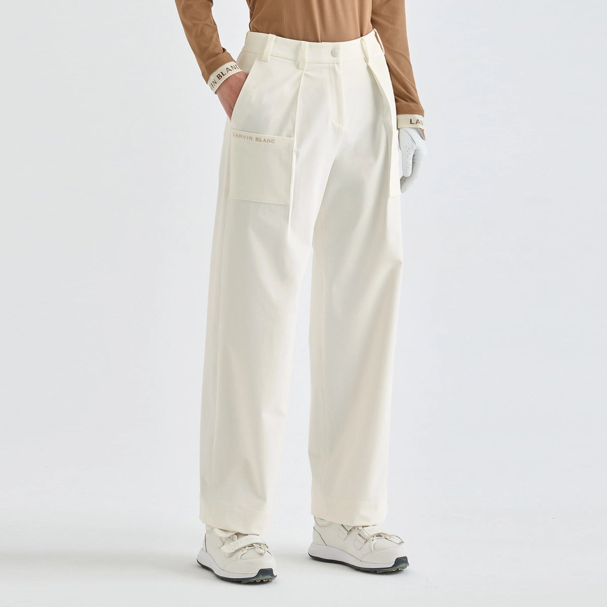 A model is shown from the waist down wearing high-waisted, wide-leg cream cargo pants with a flap pocket on the left leg, styled with a tan long-sleeve top, white gloves, and white sneakers against a plain white studio background.