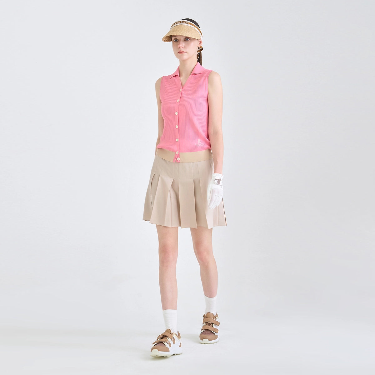 A model stands against a light gray studio background wearing a pink sleeveless button-front top, beige pleated skirt, beige visor, white gloves, white socks, and tan sandals.