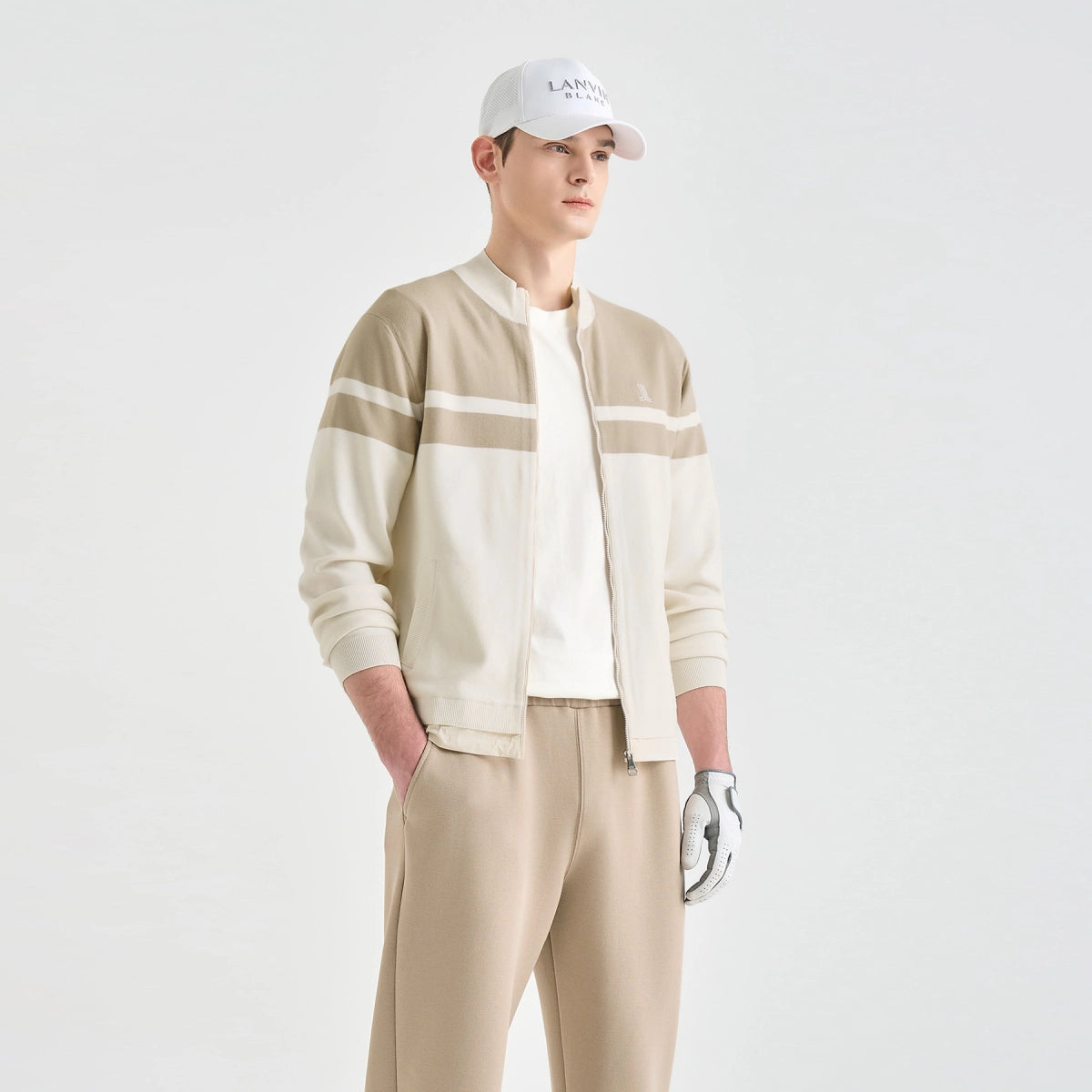 A man modeling a beige and cream color-blocked zip-up jacket, matching beige trousers, a white cap, and a white golf glove against a light gray studio background.