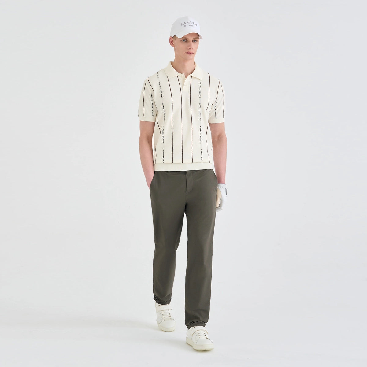 A male model stands against a light gray studio background wearing a cream short-sleeve polo shirt with vertical black stripes, olive green trousers, a white cap, and white sneakers.