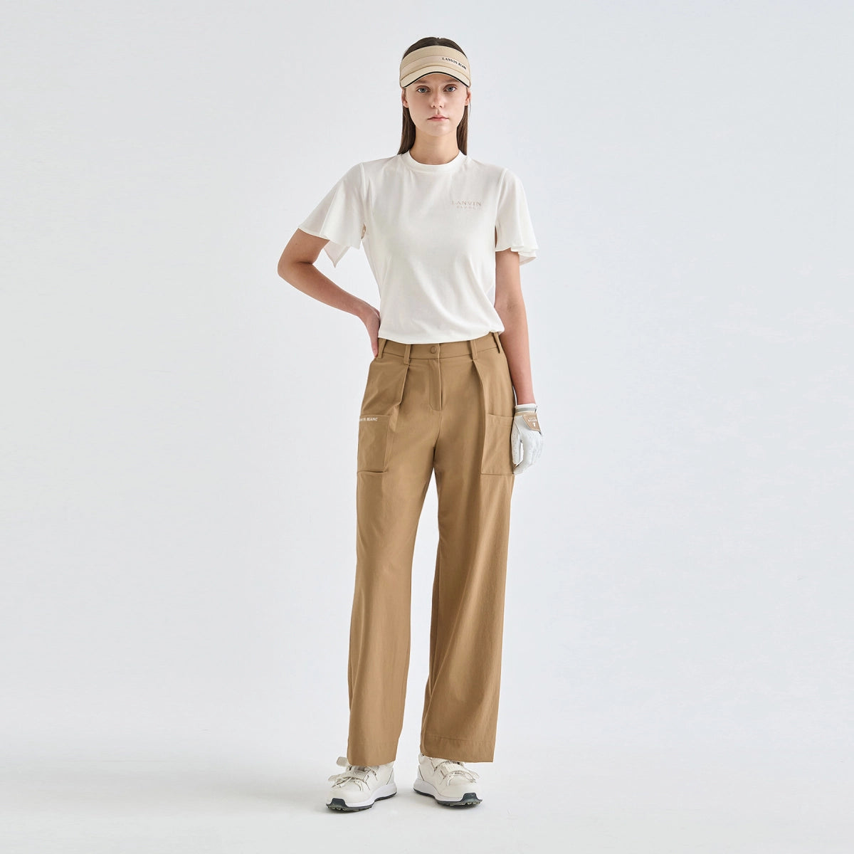 A woman modeling athletic wear consisting of a white short-sleeve t-shirt, beige wide-leg pants, a beige visor, and white golf shoes against a light gray studio background.