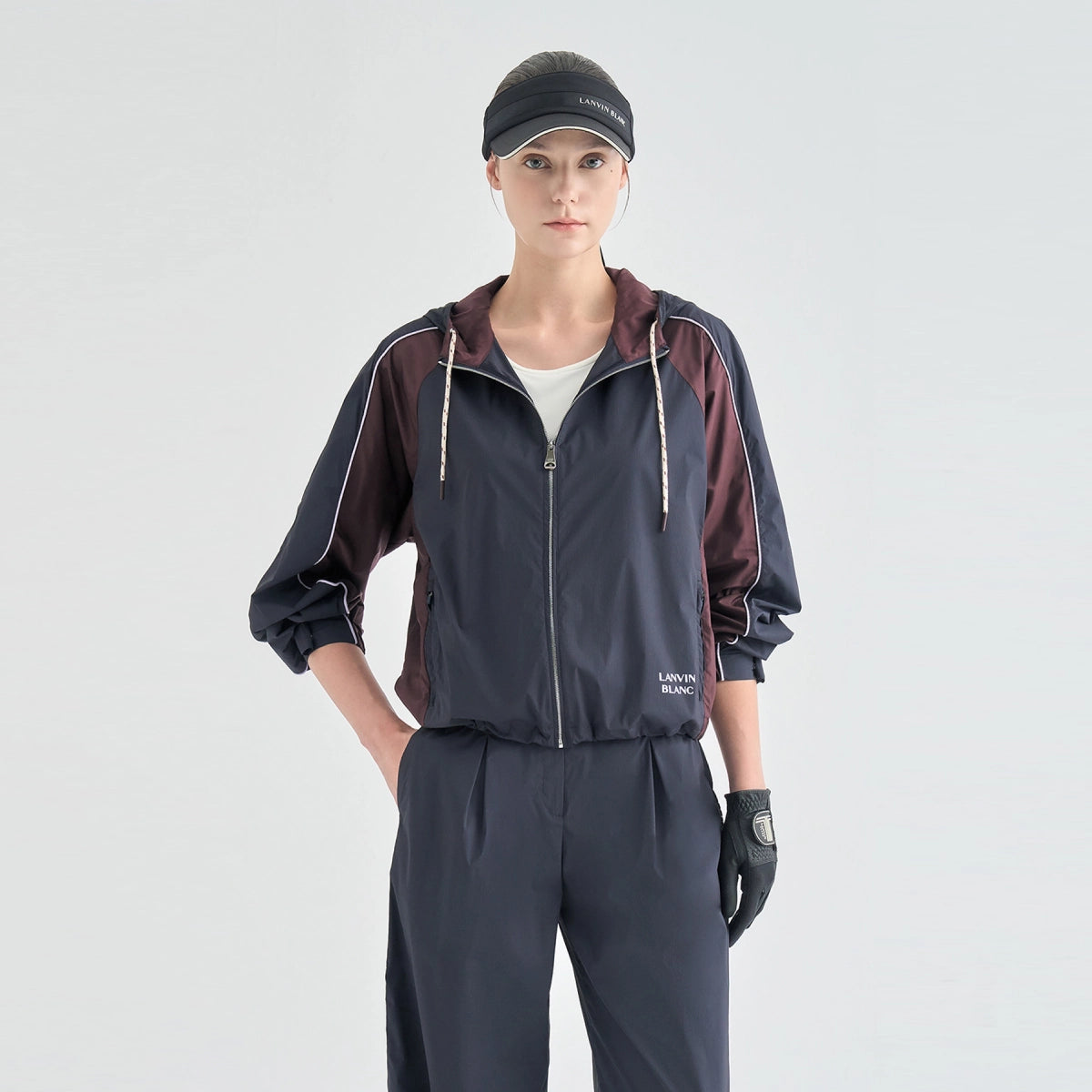 A model wearing a dark navy and burgundy color-blocked windbreaker jacket with white piping, matching dark navy joggers, a black visor, and a black glove, photographed against a light gray studio background.