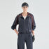A model wearing a dark navy and burgundy color-blocked windbreaker jacket with white piping, matching dark navy joggers, a black visor, and a black glove, photographed against a light gray studio background.