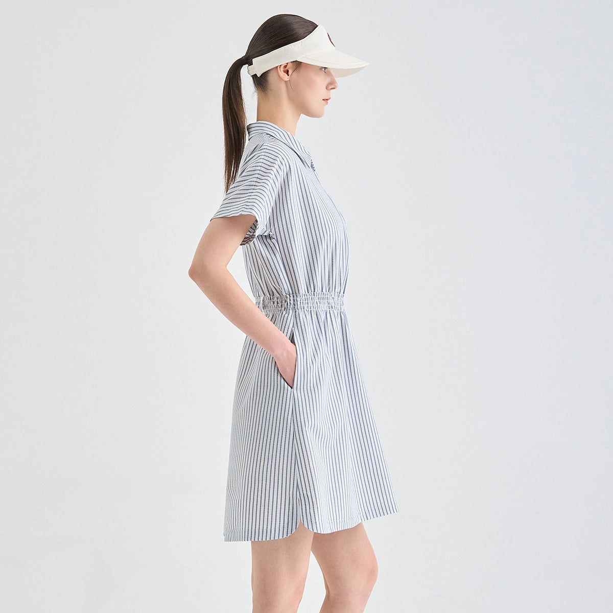 A woman in profile wearing a short-sleeve collared dress with thin vertical blue and white stripes, paired with a white visor, against a plain light gray studio background.
