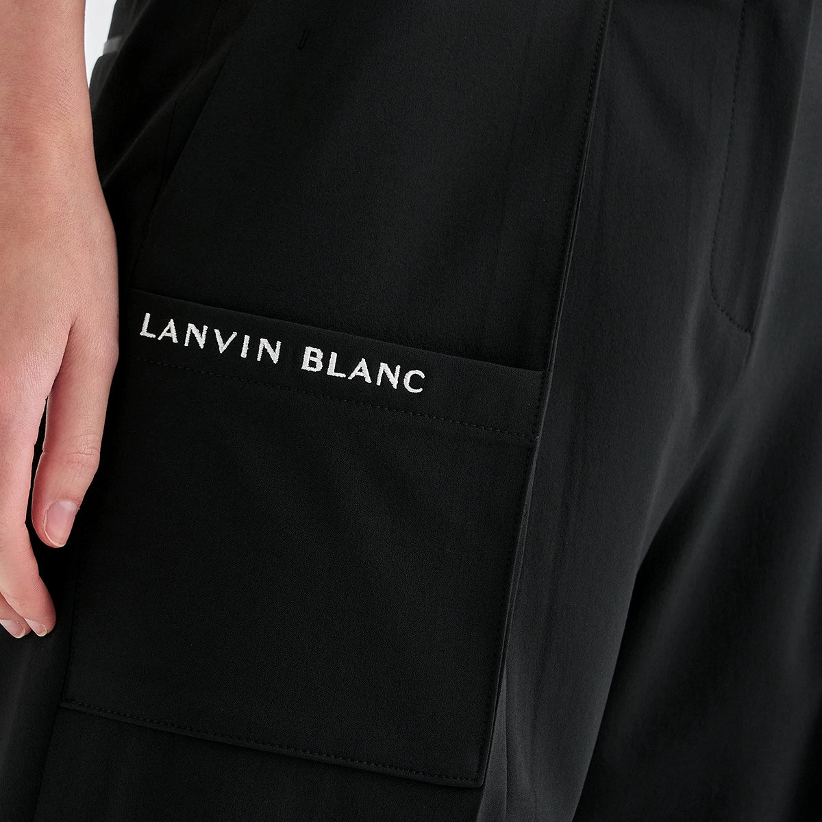 Close-up of a black garment with a front pocket featuring the white embroidered text 'LANVIN BLANC', with a hand holding the pocket edge.