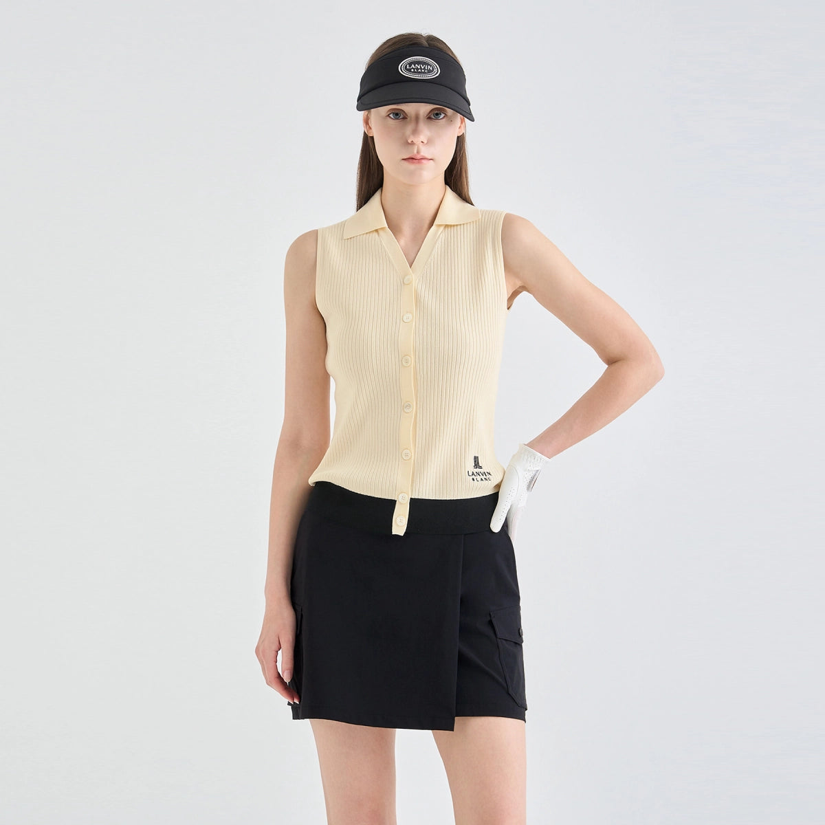 A model wearing a light yellow ribbed sleeveless collared button-up top, a black golf skirt, a black visor, and a white golf glove, photographed against a light gray studio background.