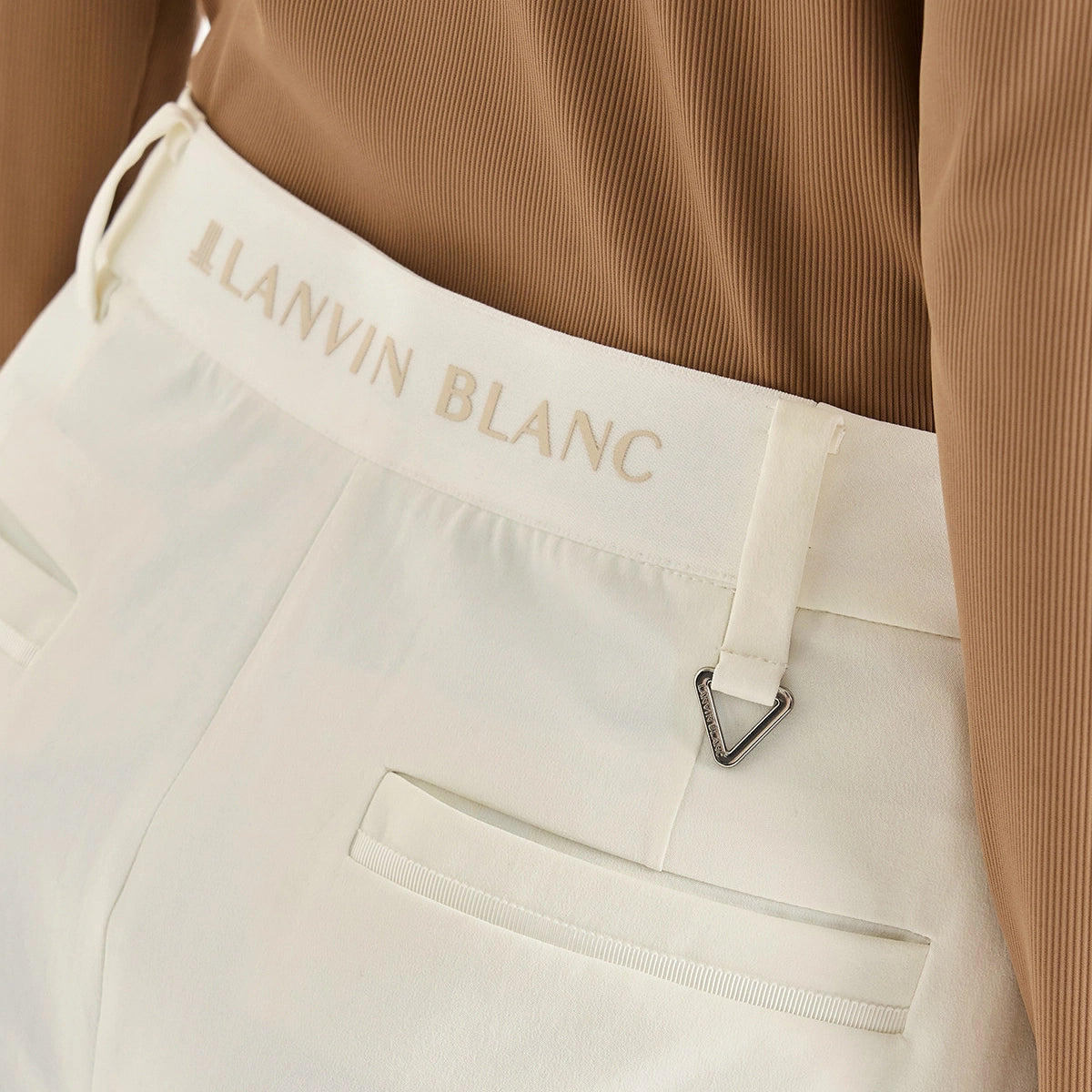 Close-up of a person wearing white Lanvin Blanc trousers with a tan ribbed top. The waistband features the 'LANVIN BLANC' logo in tan, and a small silver triangle metal accent is visible near the belt loop.