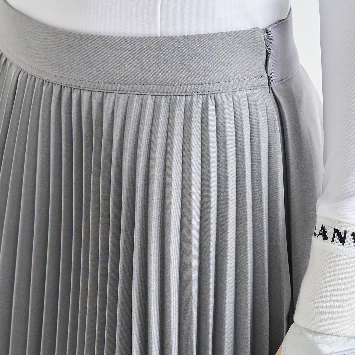 Close-up of a light gray pleated skirt with a wide waistband, shown on a white mannequin torso with a white sleeve featuring black lettering partially visible.