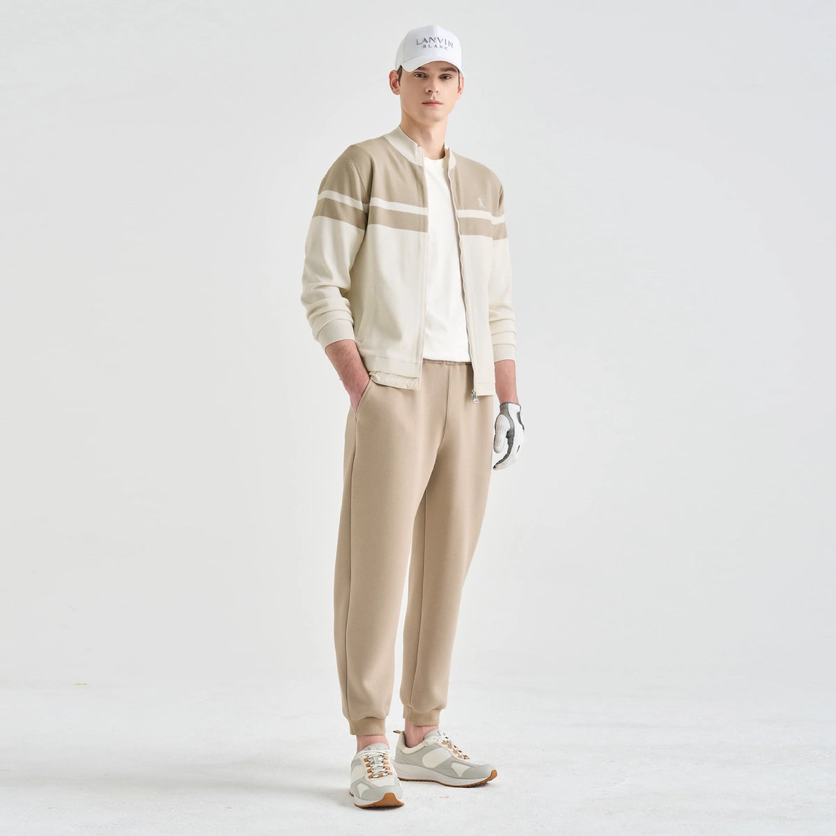 A male model stands against a clean white studio background wearing a beige and off-white striped zip-up jacket, matching beige jogger pants, a white cap, a white golf glove, and light-colored athletic sneakers.