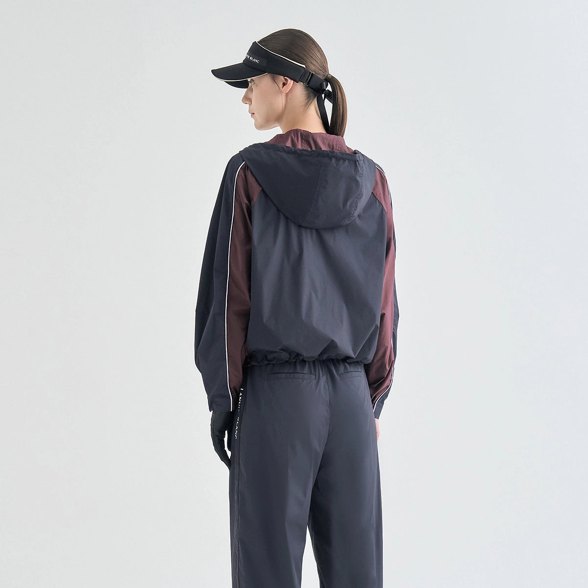 A woman shown from the back wearing a dark gray and burgundy color-blocked hooded windbreaker jacket, matching dark gray joggers, black gloves, and a black visor against a light gray studio background.