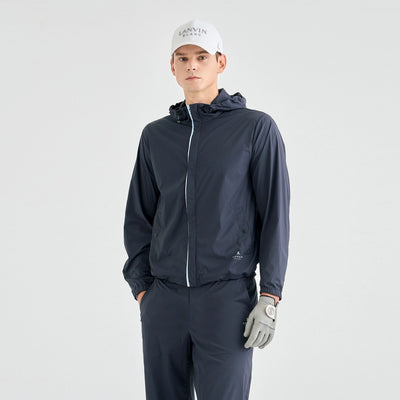 A man modeling a dark navy lightweight hooded jacket with a white zipper, matching navy pants, a white cap, and a gray golf glove, set against a plain light gray studio background.