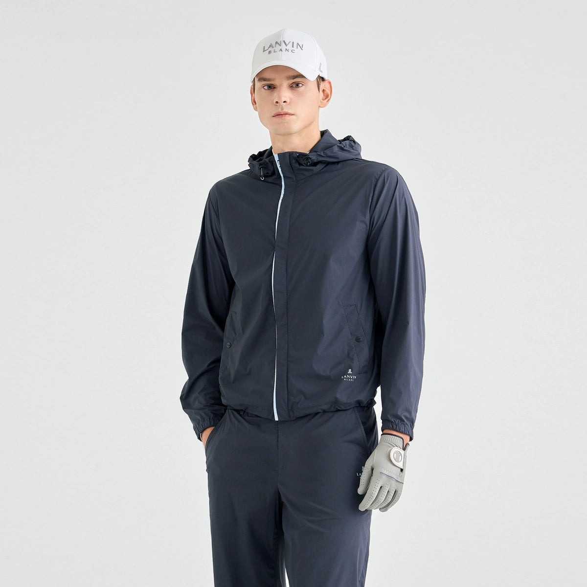 A man modeling a dark navy lightweight hooded jacket with a white zipper, matching navy pants, a white cap, and a gray golf glove, set against a plain light gray studio background.
