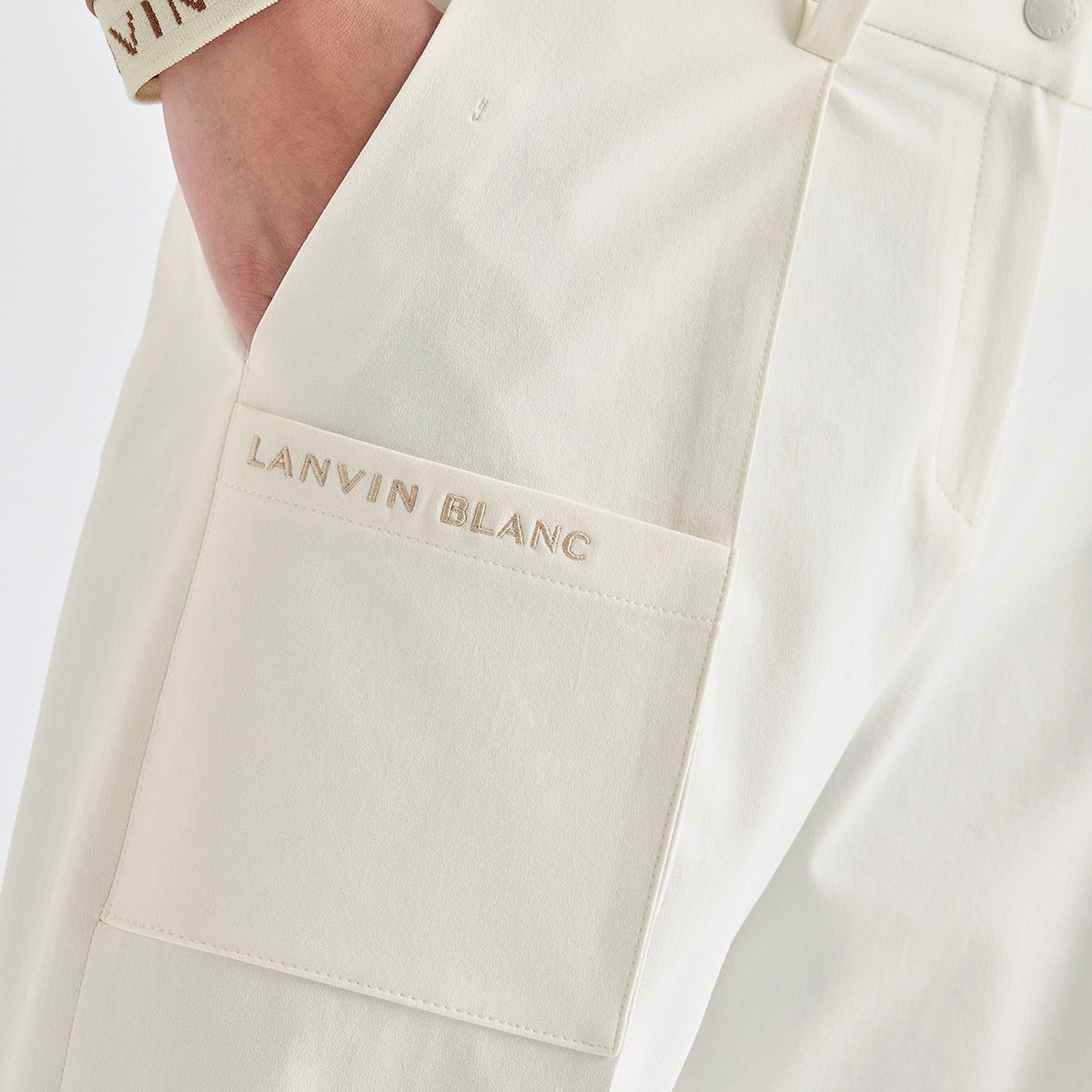 Close-up of a person wearing off-white trousers with a front patch pocket featuring the embroidered text 'LANVIN BLANC' in a matching light tone.