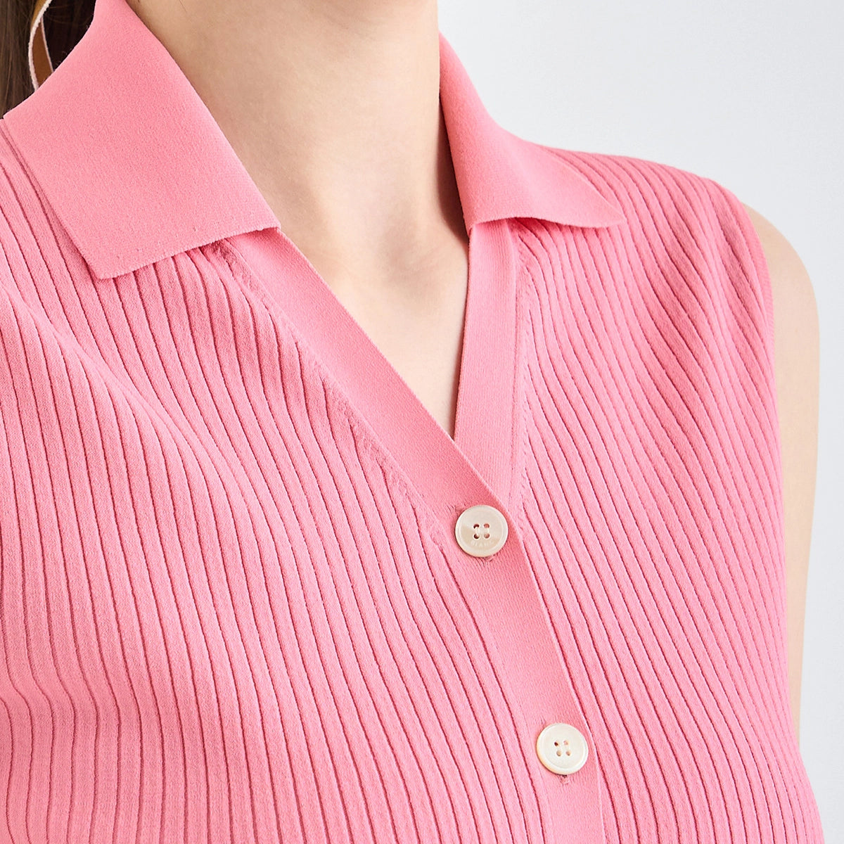 A close-up of a woman wearing a pink ribbed knit sleeveless collared top with front buttons, cropped from the neck to the upper torso against a light neutral background.