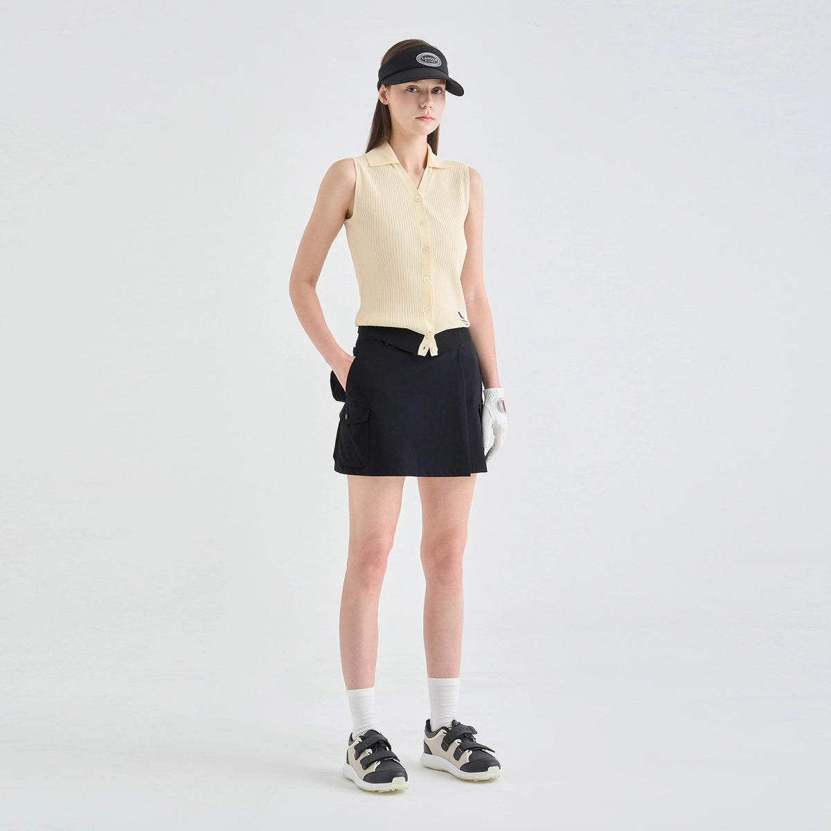 A model stands against a light gray studio background wearing a sleeveless cream polo-style top, a black skirt, a black cap, white gloves, white socks, and black-and-white sneakers.