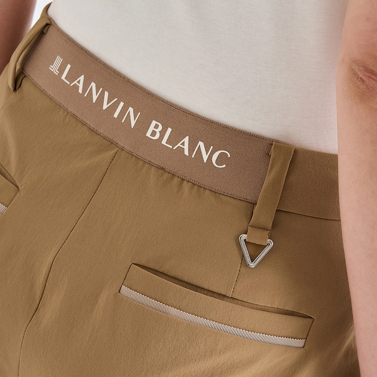 Close-up of a person wearing tan trousers with a brown waistband that reads LANVIN BLANC in white text, paired with a light cream top.