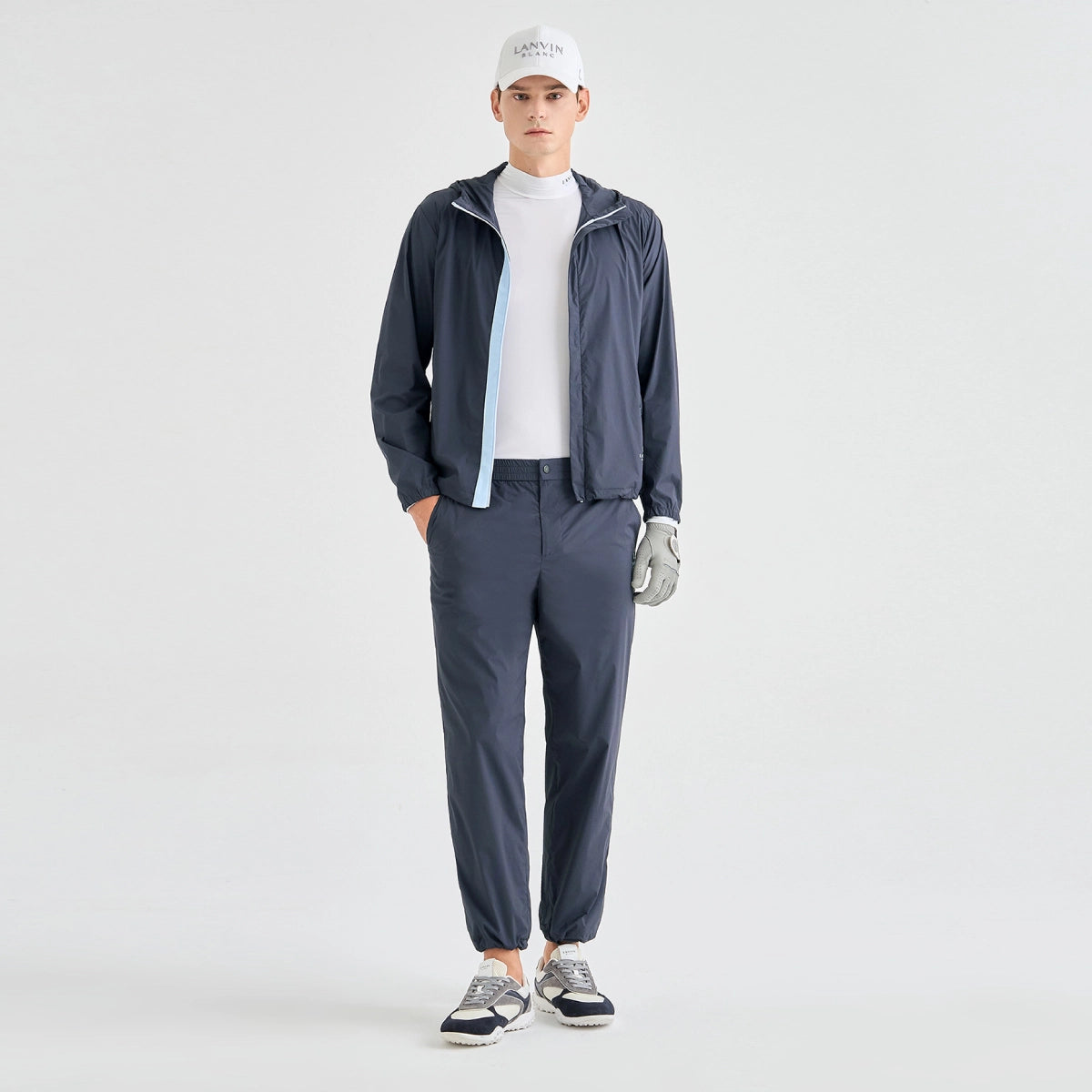 A male model stands against a light gray studio background wearing a dark navy lightweight zip-up jacket over a white mock neck, matching navy jogger pants, a white cap, and gray-and-navy sneakers.