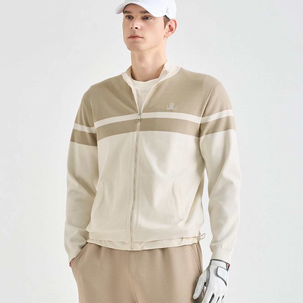 A man wearing a beige and off-white color-blocked zip-up golf jacket, beige pants, a white cap, and a white golf glove, photographed against a clean light gray studio background.