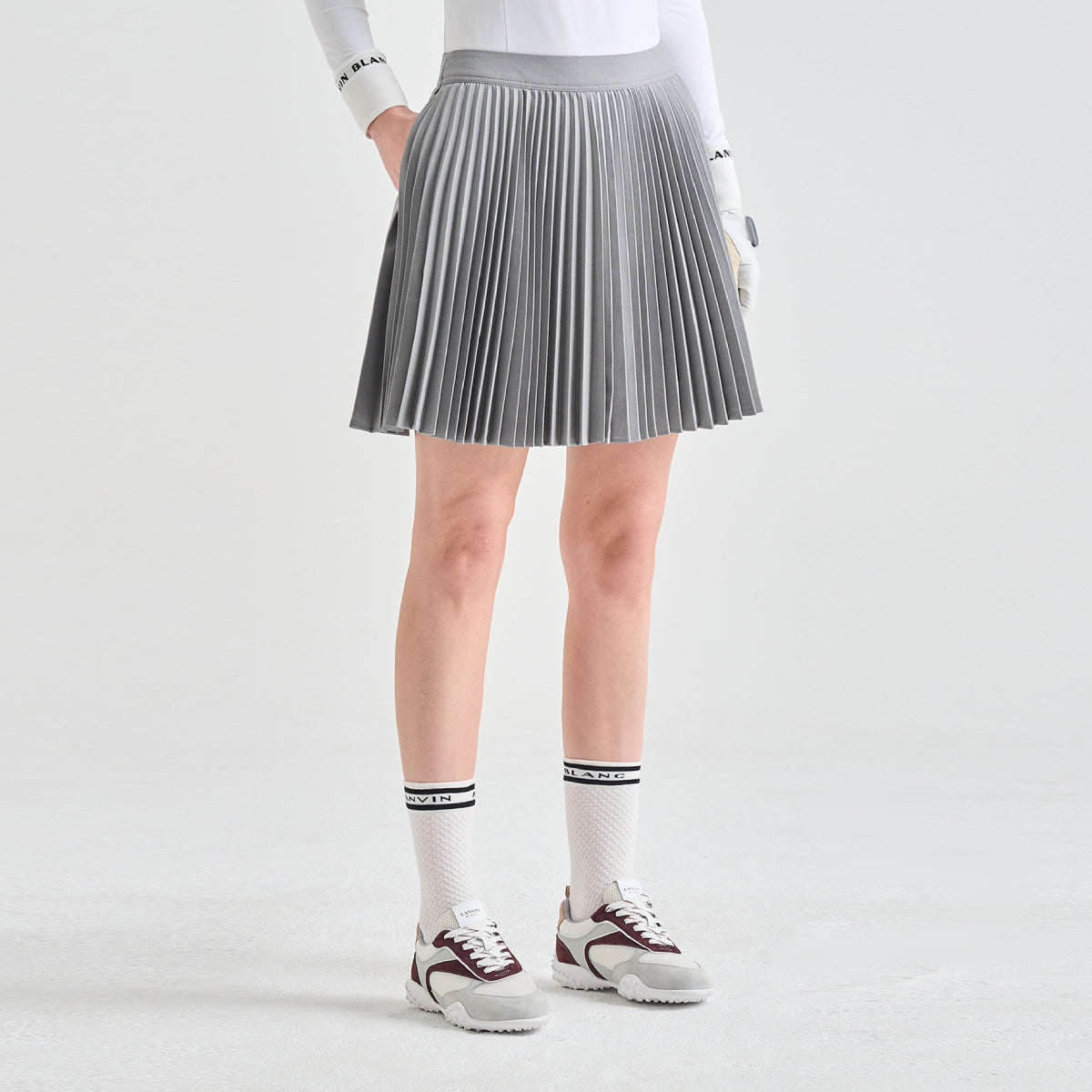 A model from the waist down wearing a gray pleated athletic skirt, white long-sleeve top, white gloves, white crew socks, and white sneakers with burgundy accents, set against a clean light gray studio background.