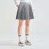 A model from the waist down wearing a gray pleated athletic skirt, white long-sleeve top, white gloves, white crew socks, and white sneakers with burgundy accents, set against a clean light gray studio background.