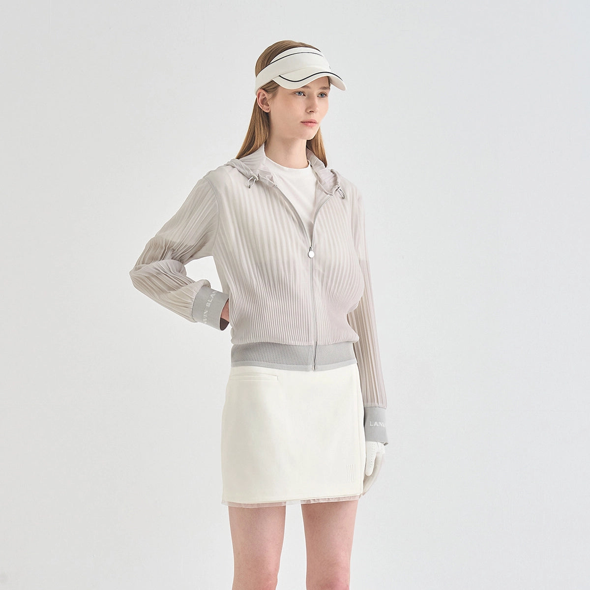 A model wearing a light beige ribbed zip-up hooded jacket with gray cuffs and hem, a white tennis skirt, a white visor, and white gloves, photographed against a solid light gray studio background.