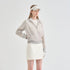 A model wearing a light beige ribbed zip-up hooded jacket with gray cuffs and hem, a white tennis skirt, a white visor, and white gloves, photographed against a solid light gray studio background.