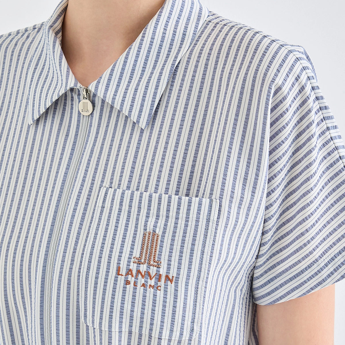 Close-up of a person wearing a short-sleeve collared shirt with narrow vertical blue and white stripes. The shirt has a chest pocket with an embroidered Lanvin Jeans logo in orange-brown on the left chest.