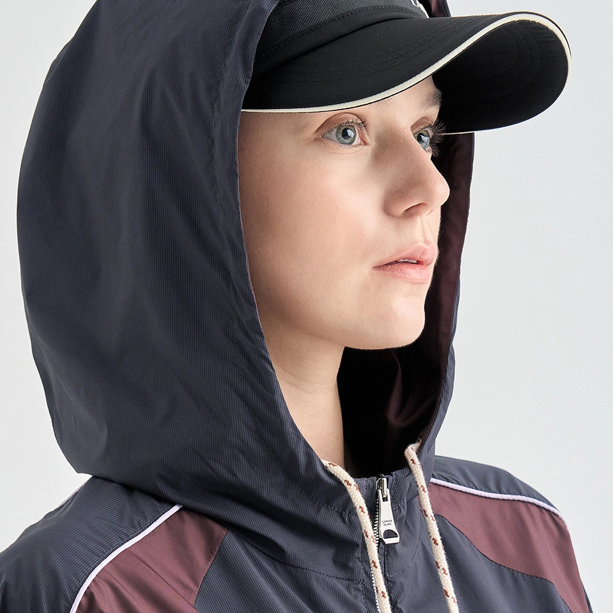 A close-up of a woman wearing a dark navy hooded windbreaker jacket with burgundy color blocking and white piping, along with a black baseball cap, against a light gray background.