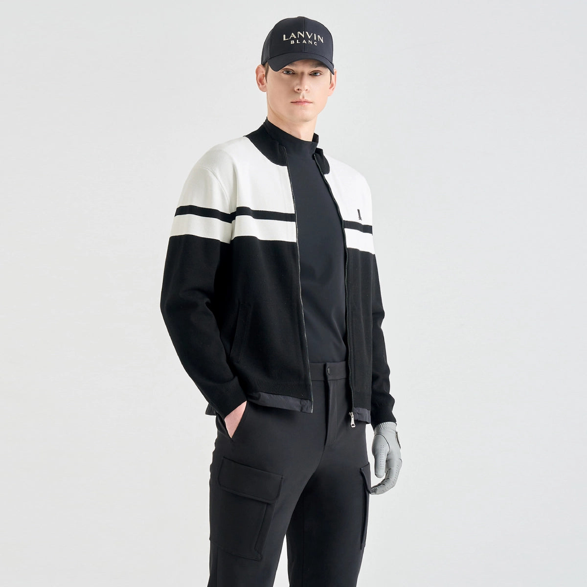 A man modeling a black and white color-blocked zip-up jacket, black pants, a black cap with 'LANVIN BLANC' text, and gray gloves, set against a plain light gray studio background.