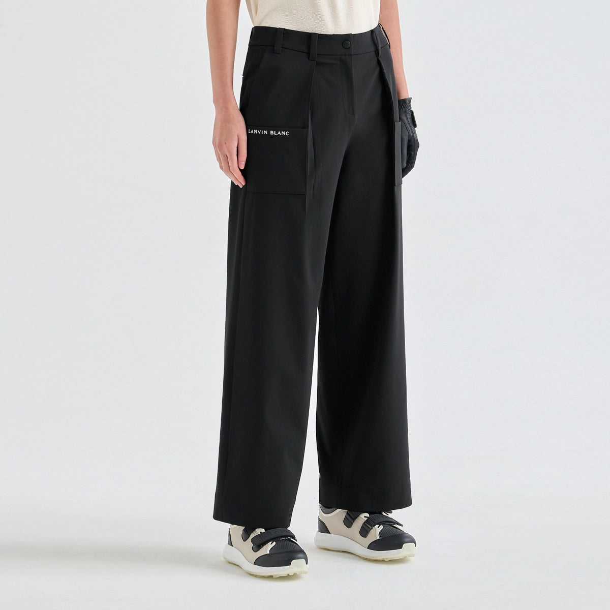 A model from the waist down wearing black wide-leg trousers with a small white 'LANVIN BLANC' logo on the left pocket, styled with a cream top and black-and-white sneakers against a light gray studio background.