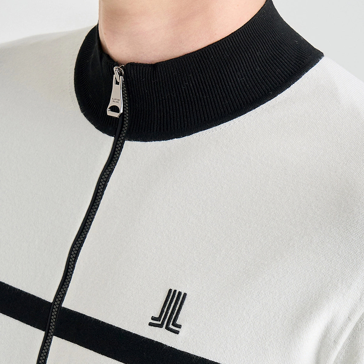 Close-up of a person wearing a white zip-up sweater with a black ribbed mock neck collar, black horizontal stripe, and a black embroidered logo on the chest.