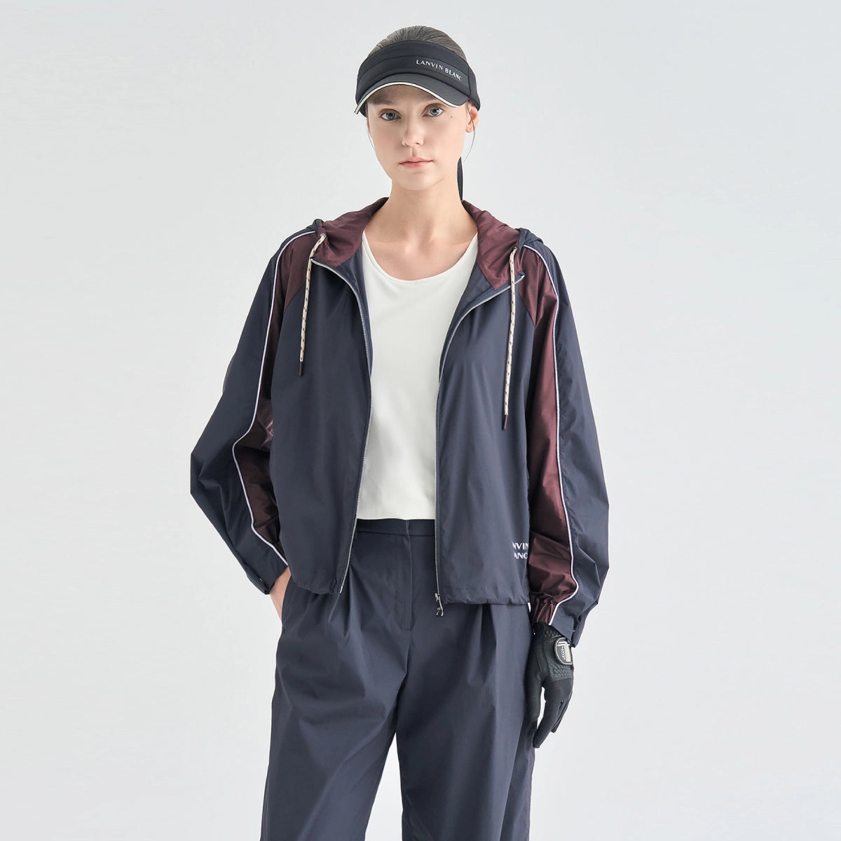 A model wearing a dark navy blue hooded windbreaker jacket with burgundy side panels and white piping, paired with matching navy pants, a white t-shirt, black gloves, and a black visor against a light gray studio background.