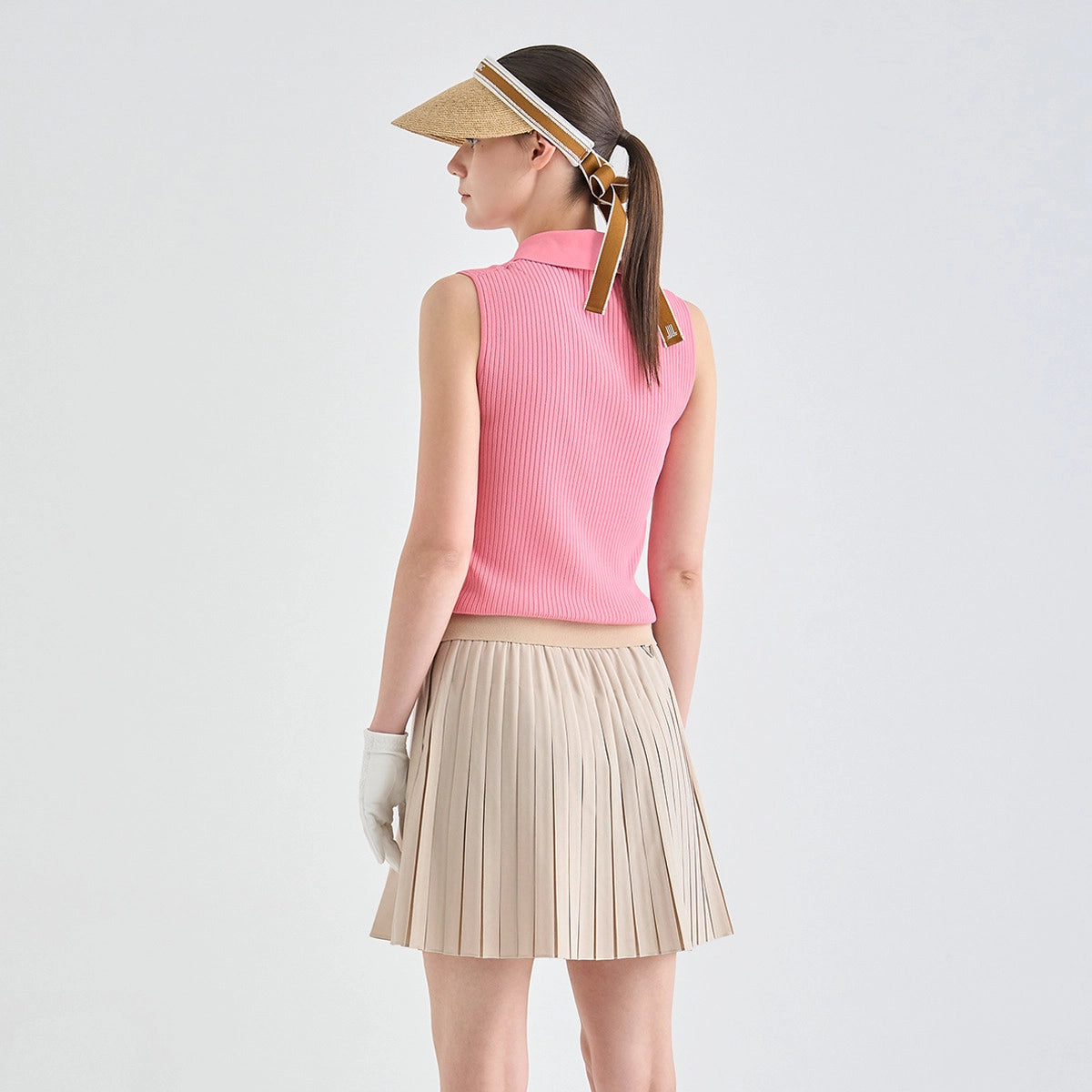 A woman shown from the back wearing a sleeveless pink ribbed knit mock-neck top, a pleated beige skirt, a straw visor, and a white glove, against a plain light gray studio background.