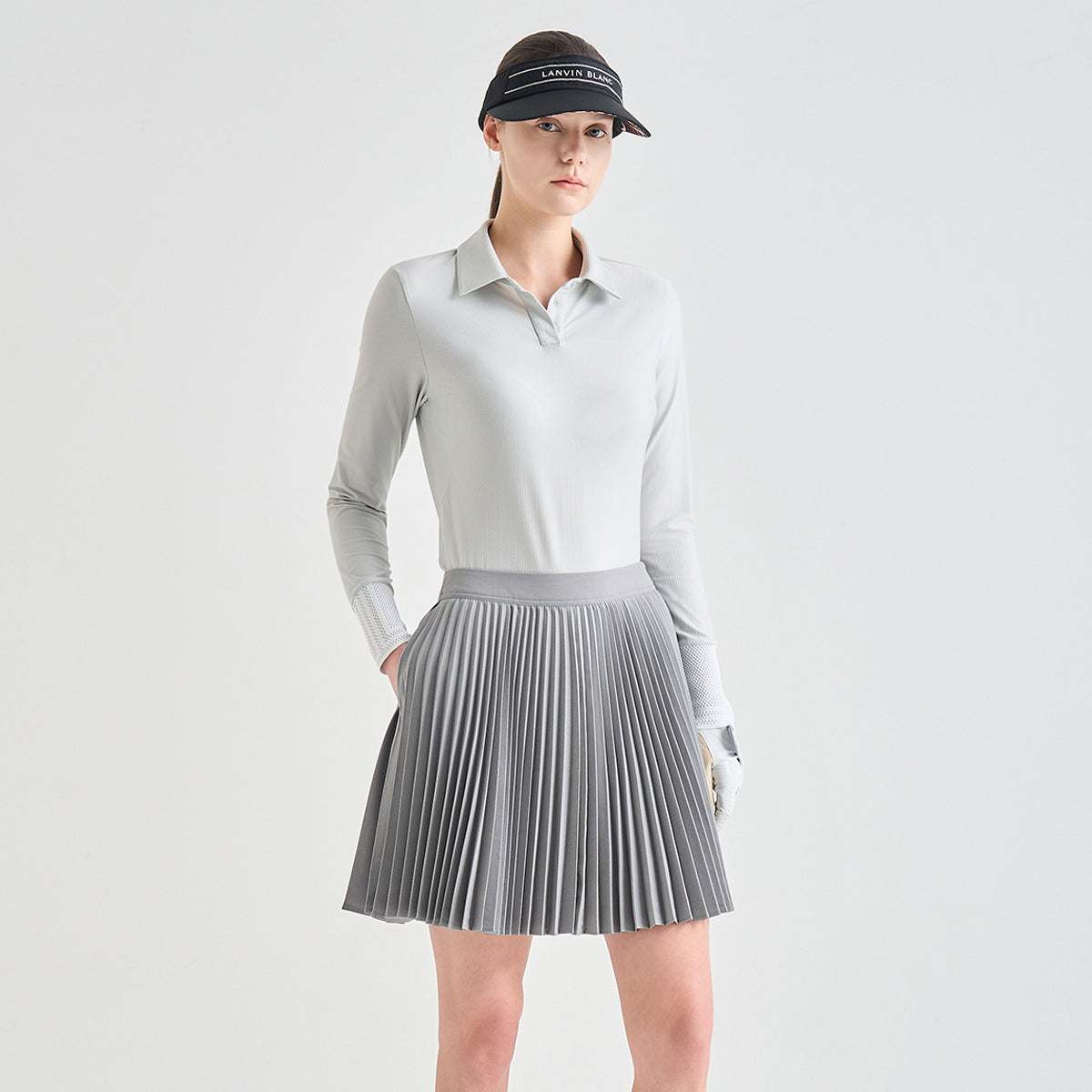 A model wearing a light gray long-sleeve collared golf shirt and a matching gray pleated golf skirt, paired with a black visor and white glove, against a light gray studio background.