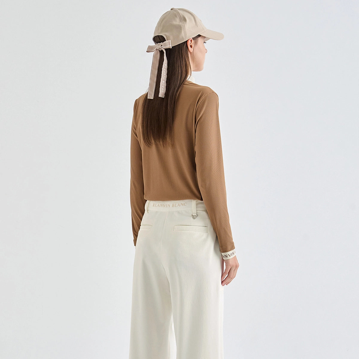 A woman shown from the back wearing a fitted brown long-sleeve top, off-white wide-leg trousers, a beige baseball cap with a ribbon tie, and a bracelet, against a plain light gray studio background.