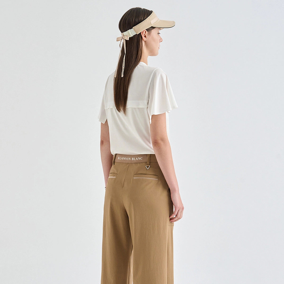 A woman shown from the back wearing a cream short-sleeve top, tan wide-leg trousers, and a beige visor against a light gray studio background.